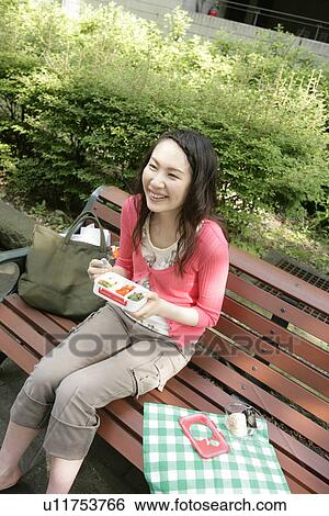 Stock Photograph - A young woman has her snacks from her lunch box while sitting on the bench. Fotosearch