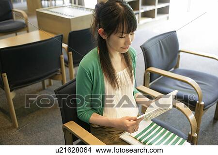 Picture - A young woman sits on the chair while she reads a book in the library. Fotosearch