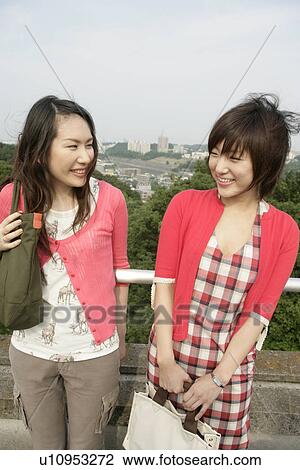 Stock Image - Two young women heartily share laughter as they stand against the railing. Fotosearch