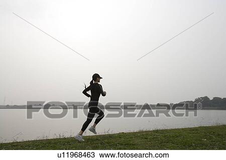 Stock Image - View of a young woman jogging beside a river. Fotosearch