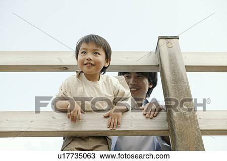 Stock Image - A small boy peeping out of wooden railing as his father looks on. Fotosearch