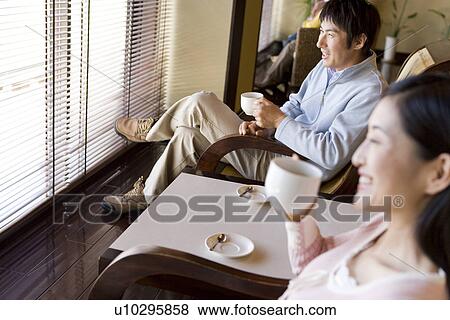 Stock Photo - Couple sitting beside the window, holding coffee cups, smiling. Fotosearch