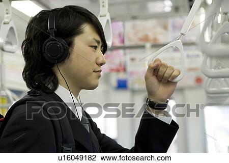 Stock Image - Young man hanging on to strap, wearing headphones. Fotosearch