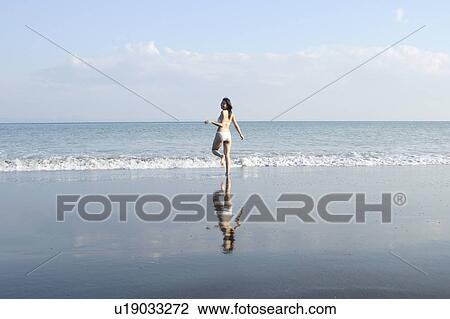 Stock Image - A young woman in bikini running at beach, rear view. Fotosearch