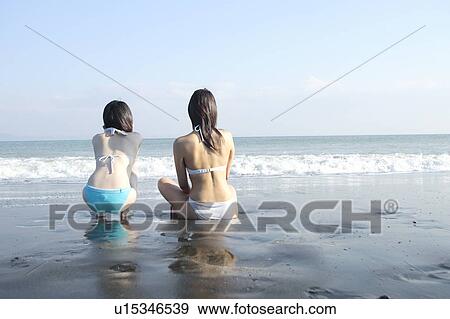 Stock Photo - Two young women in bikini sitting at beach, rear view. Fotosearch