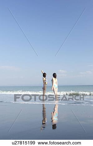 Stock Photo - Two young women in bikini standing at beach, rear view. Fotosearch