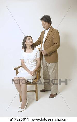 Stock Image - Mature woman sitting in chair and mature man standing. Fotosearch