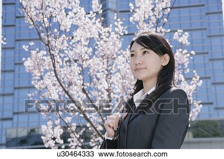 Stock Image - Young woman in suit standing in front of cherry blossoms. Fotosearch