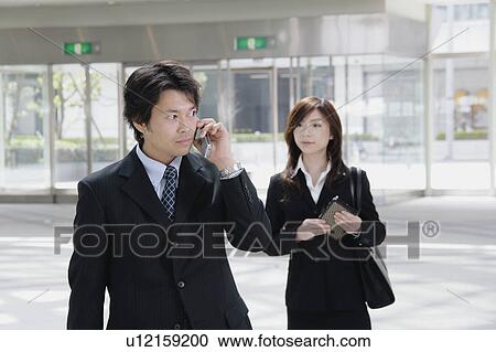 Stock Image - A mid adult man talking over mobile phone and a young woman standing behind. Fotosearch