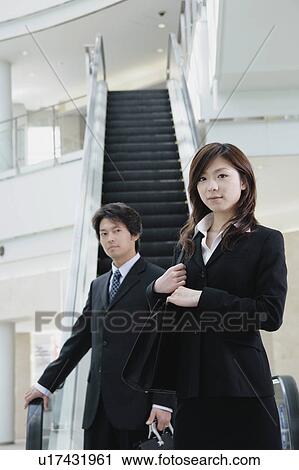 Stock Image - A young businesswoman and mid adult businessman standing in front of escalator. Fotosearch