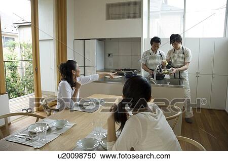 Stock Image - Father and son cooking in kitchen while mother and daughter waiting. Fotosearch