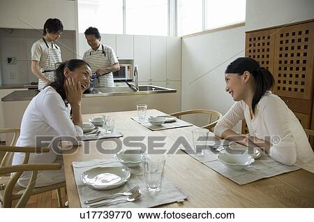 Stock Image - Mother and daughter cooking in kitchen while father and brother waiting. Fotosearch
