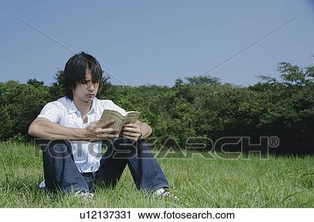 Stock Image - A young man reading a book on grass field. Fotosearch