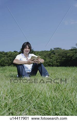 Stock Image - A young man reading a book on grass field. Fotosearch