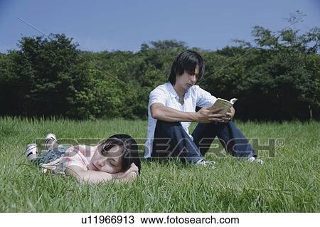 Stock Image - A young woman sleeping on grass field and a man reading a book. Fotosearch