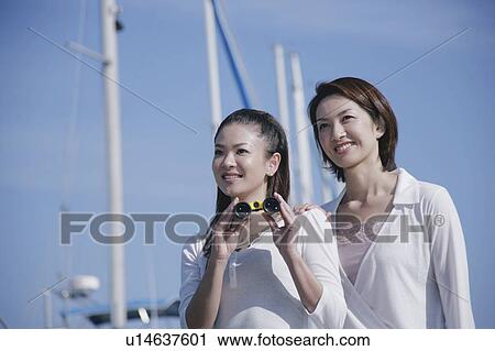Stock Image - Two young women in marina, one with binoculars. Fotosearch