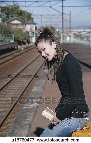 Stock Photo - Young woman sitting on suitcase on train platform. Fotosearch