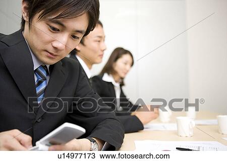 Stock Image - Young businessman using mobile phone during meeting. Fotosearch