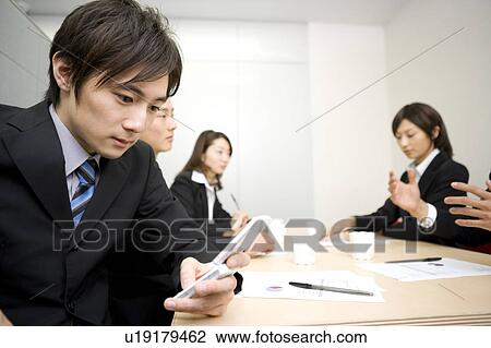 Stock Image - Young businessman using mobile phone during meeting. Fotosearch