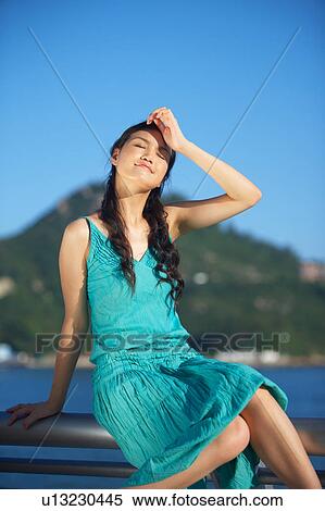 Stock Photography - Young Lady Sitting on Railing of Fence, Hand on Forehead, Eyes Closed. Fotosearch