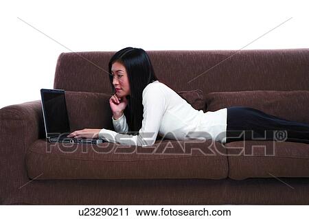 Stock Image - Young woman lying on sofa, using laptop. Fotosearch