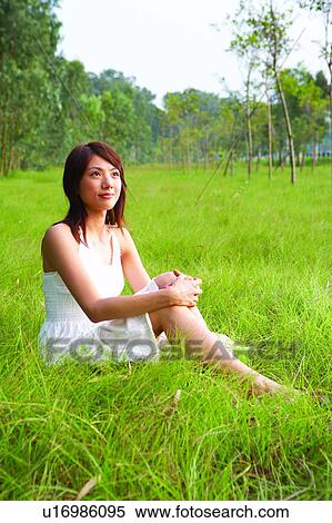 Stock Photography - Young woman sitting on grass, holding knees, full length. Fotosearch