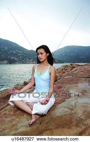Stock Photo - Young woman sitting on rock, near the sea, in yoga posture. Fotosearch