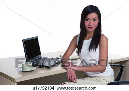 Picture - Young woman sitting, looking at camera, in office environment. Fotosearch