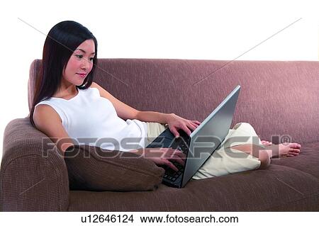 Picture - Young woman sitting on sofa and looking at laptop. Fotosearch