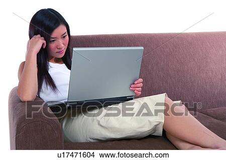 Picture - Young woman sitting on sofa and looking at computer. Fotosearch