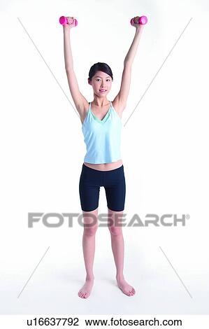Stock Image - Young woman holding dumbbell, arms raised, front view. Fotosearch