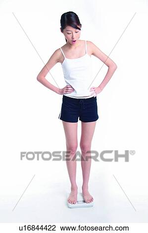 Stock Image - Young woman standing on weight scale, full length, hands on waist. Fotosearch