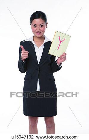Stock Image - Young woman in business suit, standing, holding a notebook with "Y" marked on, raising thumb. Fotosearch