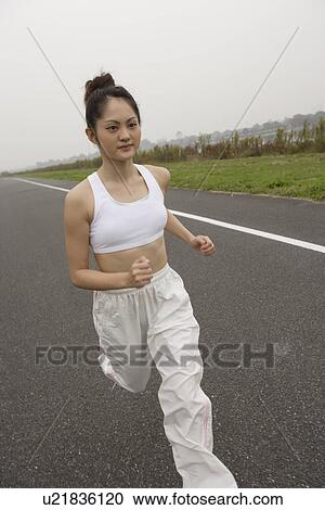 Stock Image - Side view of a young woman jogging on the road. Fotosearch
