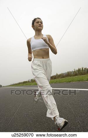 Stock Image - Side view of a young woman jogging on the road. Fotosearch