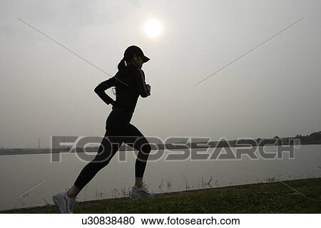 Stock Image - View of a young woman jogging beside a river. Fotosearch