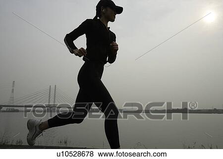 Stock Photo - View of a young woman jogging beside a river. Fotosearch