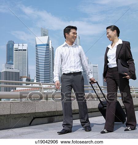 Stock Photograph - Low angle view of a businesswoman talking to a businessman and pulling a suitcase. Fotosearch