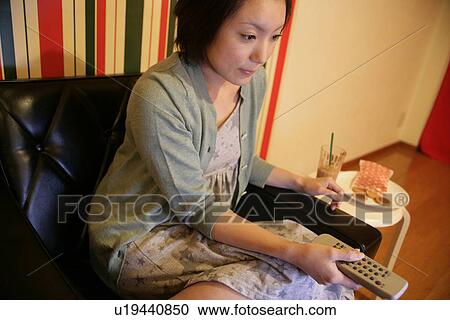 Stock Image - A young woman has her breakfast while watching television as she holds the remote control. Fotosearch