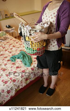 Stock Image - A young woman holds a basket full of clothes as she stands near the bed. Fotosearch