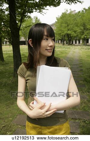 Stock Image - A young woman holds files and papers while she stands amidst grass and trees. Fotosearch