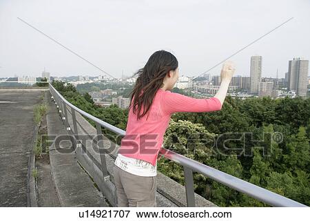 A young woman looks down and waves her hand as she stands against the railing View Large Photo Image Stock Photo - A young woman looks down and waves her hand as she stands against the railing. Fotosearch