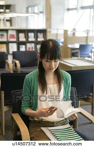 Stock Image - A young woman sits on the chair while she reads a book in the library. Fotosearch