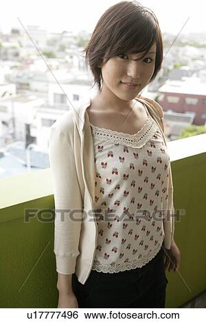 Stock Photograph - A young woman stands against the railing of the balcony as she smiles at the camera. Fotosearch