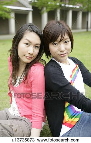 Stock Image - Two young women rest on each others back as they sit on the grassland. Fotosearch