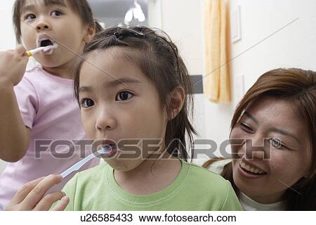 Stock Image - Two girls brushing teeth as mother watches them. Fotosearch