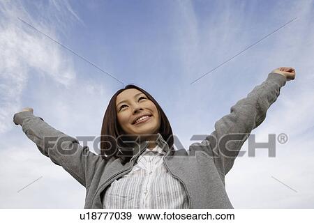 Stock Photo - View of a cheerful woman looking up at sky with her arms stretched. Fotosearch