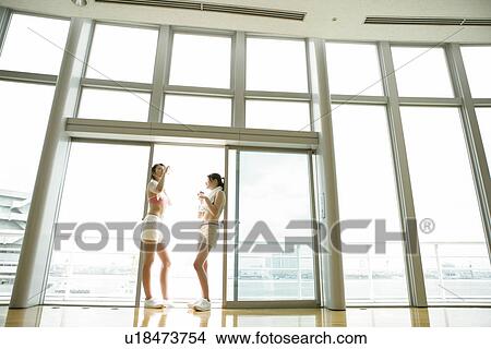 Picture - Two young women take a break from exercise as they drink water while standing near the sliding door. Fotosearch