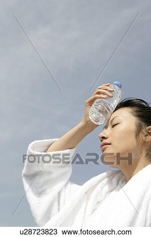 Stock Image - A woman in bathrobe placing a bottle of water on her forehead. Fotosearch