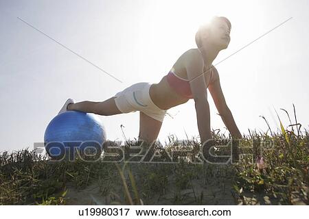 A young woman exercising under sunlight as she rests her leg on the ball with her hands touching the ground View Large Photo Image Stock Photo - A young woman exercising under sunlight as she rests her leg on the ball with her hands touching the ground. Fotosearch
